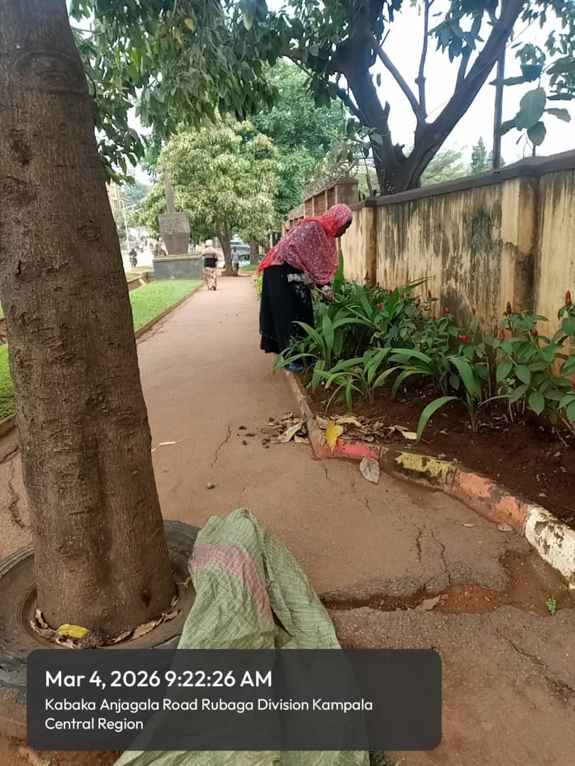 Woman in a pink headscarf and black dress tending to plants beside a weathered wall.