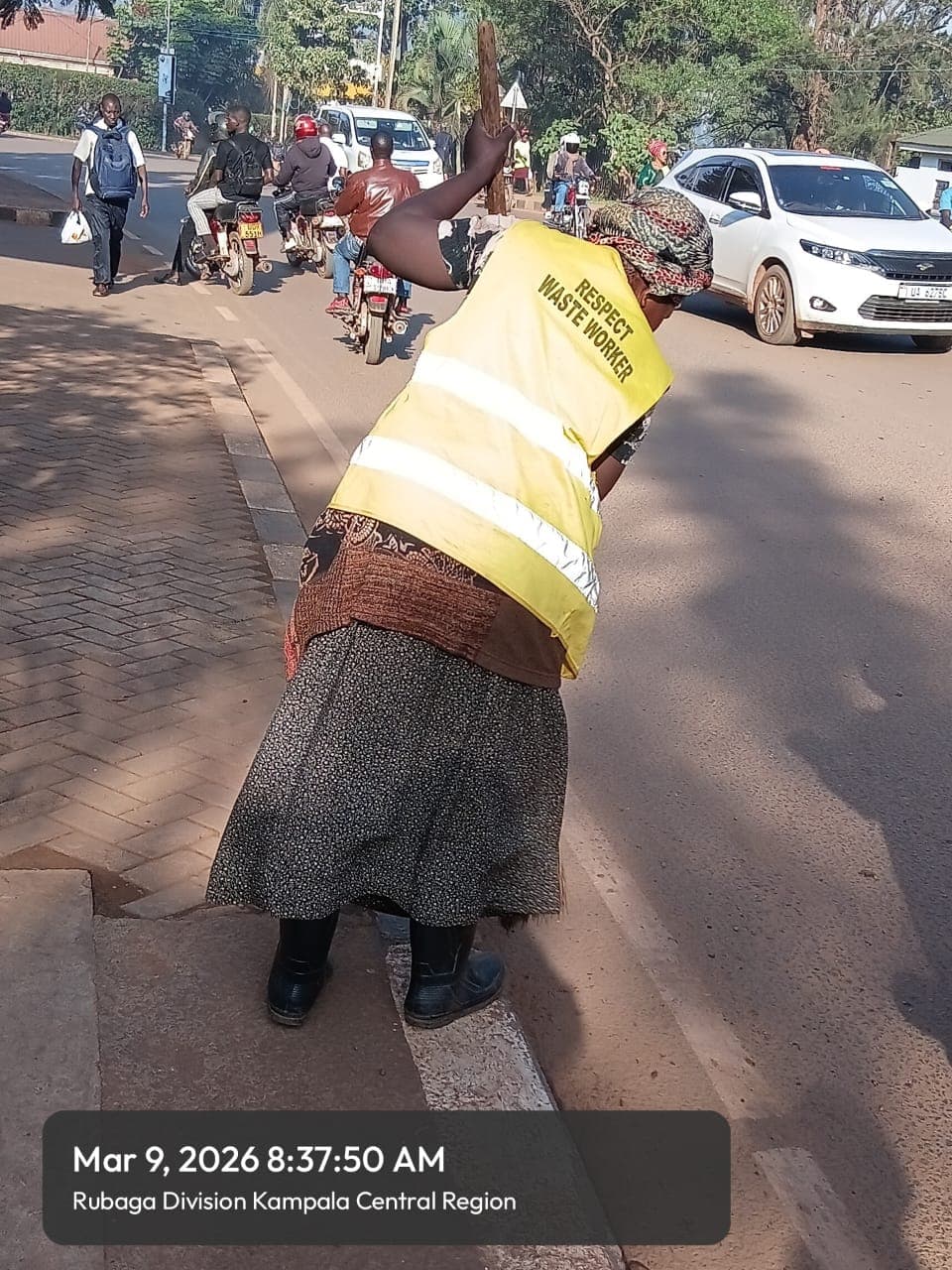 Waste worker in a yellow reflective vest sweeps a busy street in Kampala, Uganda.
