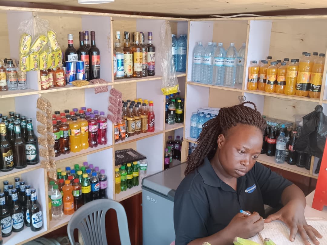 Woman with braids writing in a notebook inside a shop stocked with bottled beverages.